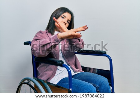 Young brunette woman sitting on wheelchair rejection expression crossing arms and palms doing negative sign, angry face 