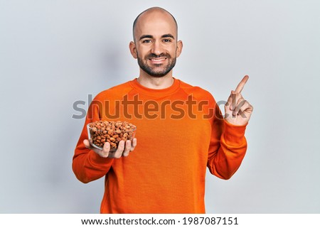 Young bald man holding peanuts smiling happy pointing with hand and finger to the side 