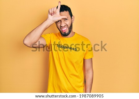 Hispanic man with beard wearing t shirt with happiness word message making fun of people with fingers on forehead doing loser gesture mocking and insulting. 