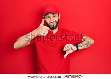 Hispanic man with beard wearing delivery uniform and cap doing thumbs up and down, disagreement and agreement expression. crazy conflict 