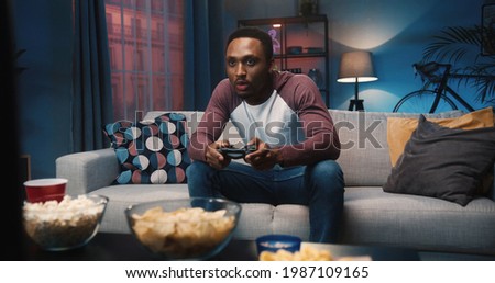 African American young joyful man resting at home sitting on couch and playing video games in evening alone. Handsome guy gamer plays game on console having fun at night. Entertainment concept