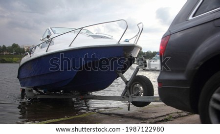 A 4x4 black car pulls blue-white trimaran cabin motor boat on trailer on concrete slipway into the river water, boat launch before active recreation on the river Royalty-Free Stock Photo #1987122980