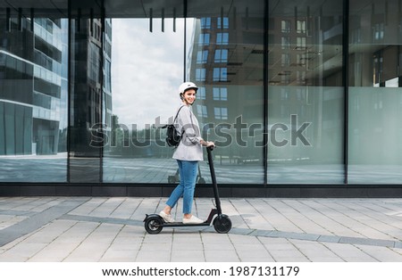 Side view of young smiling businesswoman with cycling helmet on her head driving an electrical push scooter Royalty-Free Stock Photo #1987131179