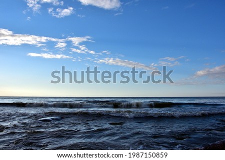 Waves on the evening sea before sunset under a blue sky with light clouds, which themselves begin to cast light shadows on the sky.