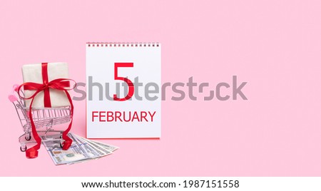A gift box in a shopping trolley, dollars and a calendar with the date of 5 february on a pink background.