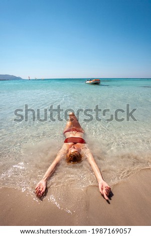 Beautiful happy woman in swimsuit laying in the shallow sea water with pink sand on Elafonissi beach on Crete island.