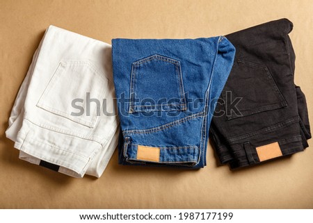 White blue black jeans pants stacked. Assortment of different color denim clothing in store shop. White denim jean pants, blue jeans, black denim. Top view on brown table Royalty-Free Stock Photo #1987177199