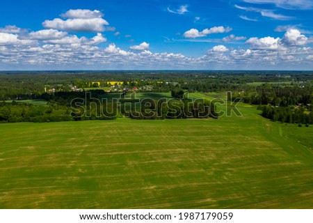 aerial view of a green  field with traces of wheels from a tractor, a dense forest grows along the edges, all under a blue sky with beautiful white clouds.