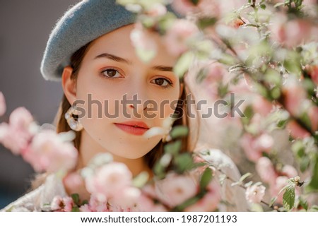 Young beautiful woman wearing classic blue beret, pearl earrings, posing in blooming garden. Spring fashion, lifestyle, concept. Copy, empty space for text.