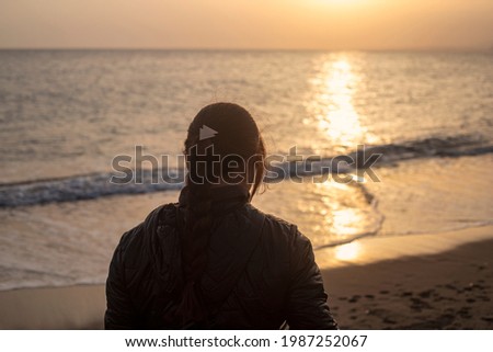 Photography of young and white woman looking at the sea during the 
sunset, with a braid and coat, during the winter.