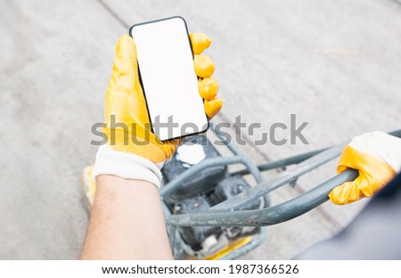 The worker holds the phone while tamping a gravel by the vibration plate. Mockup for house repair or building Royalty-Free Stock Photo #1987366526