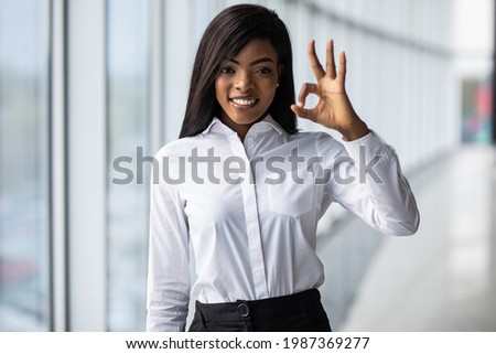 Enthusiastic african female student wears white t-shirt successfully passed test and happy for it. Indoor portrait of girl with light-brown skin posing with okay sign near to working colleagues.