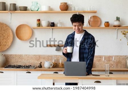 Home comfortable office, work remotely online, new normal during covid-19. Busy happy 30s young asian guy employer holding coffee cup in modern kitchen interior with laptop on table, free space