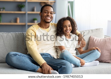 Adorable black family father and daughter watching TV together at home. Smiling african american dad and school girl sitting on couch and hugging, posing in living room, copy space