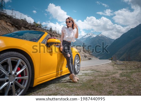 Beautiful Asian Chinese girl in sunglasses next to fancy yellow sport car posing and enjoys life during trip to mountains. Bright windy day, clear sky, fluffy clouds. Luxury travel around Sichuan.