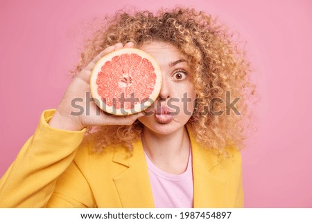 Photo of surprised woman with curly hair keeps half of grapefruit over eye folded lips keeps to healthy diet wears yellow jacket isolated over pink background. Citrus fruit for making cosmetic product