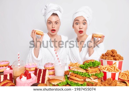 Stunned diverse women crave to eat donuts dressed in domestic clothes have temptation to eat junk food have tasty dinner at home break diet isolated over white background. Unhealthy eating concept