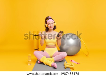 Pleased Asian sportwoman holds smartphone checks results of fitness training makes korean like sign being in good mood does yoga poses on comfortable karemat isolated over yellow background.