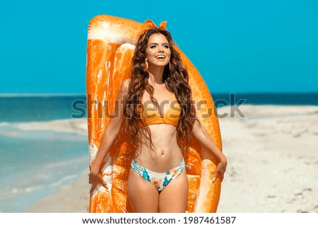 Beautiful hippie girl portrait by orange Inflatable pool float mattress on tropical beach. Fit Brunette with long healthy curly hair and makeup on summer vacation.