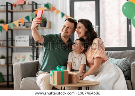 family, holidays and people concept - portrait of happy mother, father and little son with gifts and smartphone taking selfie on birthday at home party