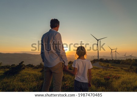 Cinematic shot of carefree young father engineer keeping his daughter for hand and looking on windmill field at sunset. Concept of renewable energy, love for nature, family, electricity, green, future Royalty-Free Stock Photo #1987470485