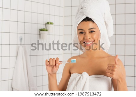 young african american woman wrapped in towels holding toothbrush and showing ok gesture in bathroom
