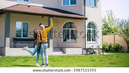 Modern beautiful excited young couple in love standing in front of the new big house with the keys in hands and hugging while they celebrating this buying Royalty-Free Stock Photo #1987515593