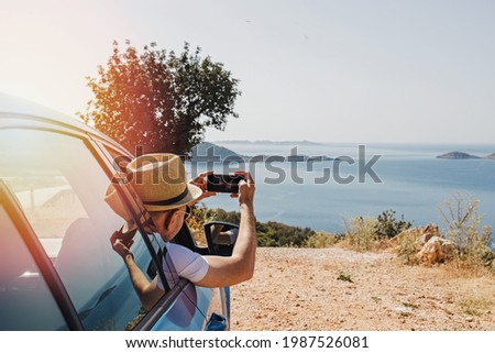 Man taking picture with smartphone from open window of a car stopped roadside with a beautiful view to the coast
