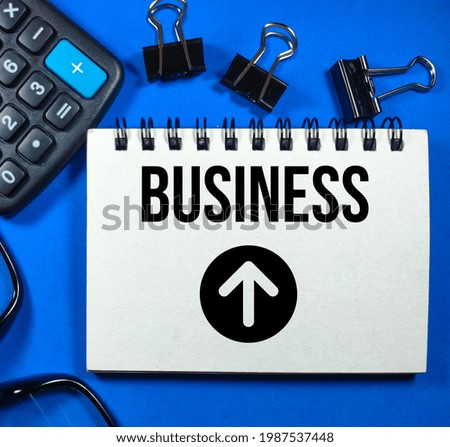 Business concept.Text Business and arrow icon on notebook with calculator,glasses and paper clips on blue background.