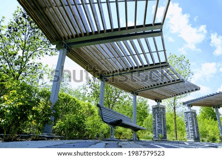A brown wooden bench with a curved back. There is a wooden canopy above it. Sky and trees in the background.
