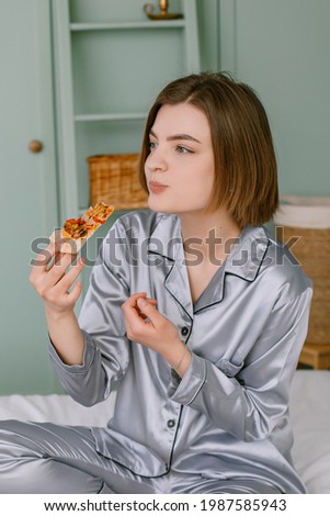 Young woman sits on the sofa eating flour pizza Interior Scandinavian style of green color Breakfast in the bed of a lonely lady Clothes gray pajamas near the phone and laptop Morning snack in bed