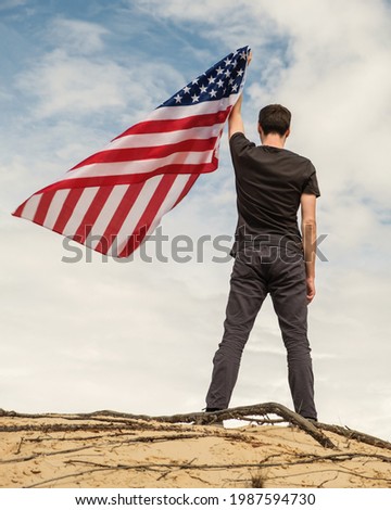 A man with an American flag stands on the sand, skies on the background. Man is holding waving American USA flag, left arm is up. Fourth of July Independence Day.