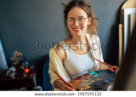 Indoor portrait of a pretty artist female sitting next to the easel with canvas painting something in her art studio. A young woman painter in transparent spectacles draws in the workshop.