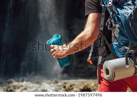 Hand close up with drinking water bottle. Man with backpack dressed in active trekking clothes touristic staying near mountain river waterfall and enjoying Nature. Traveling, trekking concept image Royalty-Free Stock Photo #1987597496