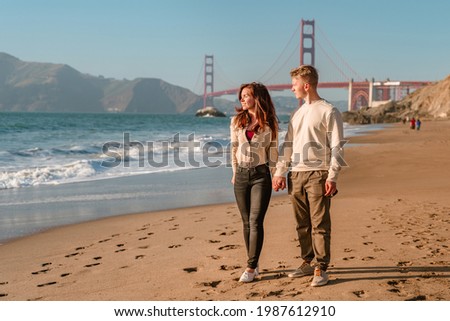 A young man and a woman take a romantic walk on the beach overlooking the Golden Gate Bridge at sunset in San Francisco