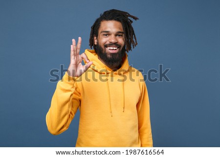 Cheerful young african american man guy wearing casual yellow streetwear hoodie posing isolated on blue wall background studio portrait. People sincere emotions lifestyle concept. Showing OK gesture