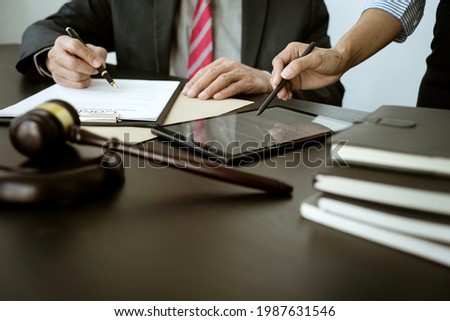 The lawyer is write drafting a legal document while listening to the assistant use her tablet to give information on the table with scale and gavel