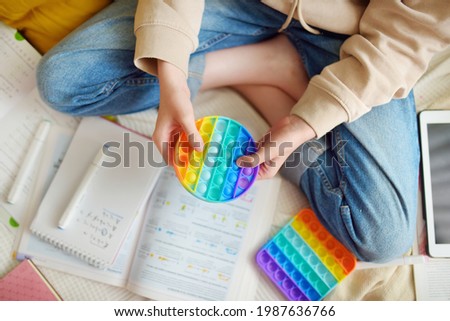 Teenage girl playing with rainbow pop-it fidget toy while studying at home. Teen kid with trendy stress and anxiety relief fidgeting game. Popping the dimples of sensory silicone toy. Royalty-Free Stock Photo #1987636766