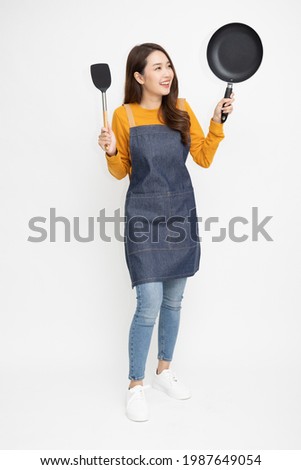 Young Asian woman housewife wearing kitchen apron cooking and holding pan and spatula isolated on white background, Full body composition Royalty-Free Stock Photo #1987649054