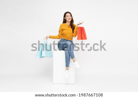 Young Asian woman holding shopping bags and sitting on white box isolated on white background Royalty-Free Stock Photo #1987667108