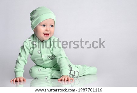 joyful infant baby kid sitting in the cotton jumpsuit with a print of the patches and the corresponding hat, and with joy and interest in looking into the corner of the frame.