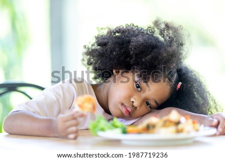 A young, curly haired African American girl sits bored looking at food. Do not want to eat food. Childhood concepts and healthy eating.