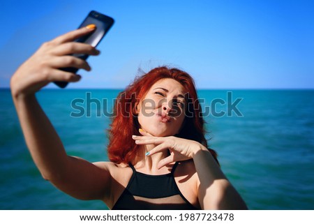 Red-haired woman squints in sun and takes selfie on smartphone camera. Lips like a duck. Sunny summer day. Cute girl takes pictures of herself against background of blue sea and sky.