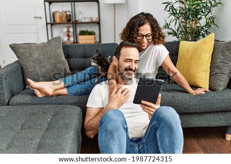 Middle age hispanic couple smiling happy and using touchpad. Sitting on the sofa with dogs at home. Royalty-Free Stock Photo #1987724315