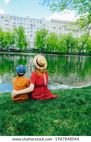 young slender mother in red dress and straw hat and her preschooler son in stylish cap sit by pond in city on green bright grass and hug. family outing. family values and embraces mother and child
