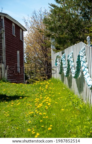 A white wooden fence with a large braided green rope hanging on the fence. The ground next to the fence is green with small yellow flowers. Behind the fence are trees and a red vintage wood building.