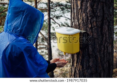 Woman in blue raincoat washes her hands in wash basin hanging on tree. Morning after rain at tourist camp in the forest by the river. Girl's face hidden by hood. Hiking lifestyle.
