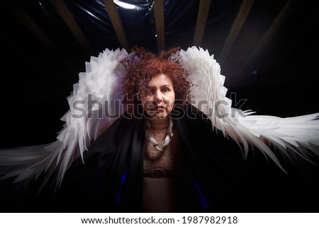 A brunette curly girl in black dress looking like an evil angel with white wings on a black background. Model and actress posing in the studio