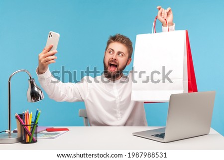 Positive bearded man blogger making selfie or recording video, posing at smartphone camera with shopping bags, bragging with purchases. Indoor studio shot isolated on blue background Royalty-Free Stock Photo #1987988531