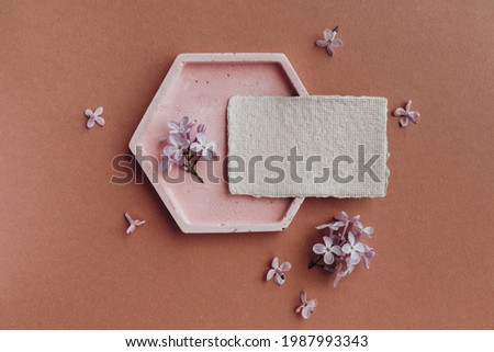 Still life scene with plate, lilac flowers and blank business, greeting card, invitation mockup on brown background. Flat lay, top view.
